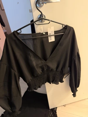 Svart  blus med v-ringning - (OANVÄND,PRISLAPP KVAR) Trendig svart croppad blus med v-ringning och ballongärmar. Toppen har smockad midja som ger en snygg siluett och är tillverkad i ett lätt, vävt material. Perfekt för dig som vill ha en stilren och modern look.