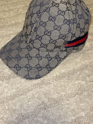Gucci keps med GG-mönster - Snygg keps från Gucci i beige och blått med klassiskt GG-monogram över hela kepsen. På sidan finns en röd och marinblå rand som ger extra lyxig känsla. Kepsen har böjd skärm och knapp på toppen. Materialet ser ut att vara canvas.