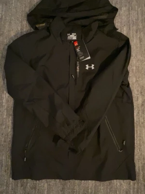 Svart vindjacka från Under Armour - Snygg svart vindjacka från Under Armour med huva och dragkedja framtill. Jackan har två fickor med dragkedja, elastiska muddar vid ärmsluten och diskret logga på bröstet. Tillverkad i lätt och tåligt material, perfekt för blåsiga dagar. Men om du ändå vill gå runt med stil.