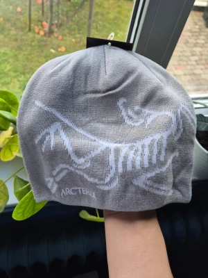 Grå mössa från Arc'teryx - Snygg grå mössa från Arc'teryx med ett stort vitt fossiltryck på framsidan. Tillverkad i mjukt stickat material som sitter skönt på huvudet. Perfekt för kalla dagar och streetwear-stil. 