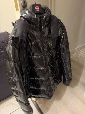Svart glansig pufferjacka Philipp Plein - Säljer en svart glansig pufferjacka från Philipp Plein med hög krage, dragkedja framtill och logotyp på bröstet. Jackan har vadderad design, avtagbar huva och flera praktiska fickor. Perfekt för kalla vinterdagar och riktigt snygg streetstil. Den är äkta har bevis om ni budar så kan jag föra fram beviset! Den är köpt på farfetch värd 20k men den ligger bara hemma priset är inte hugget i sten men jackan ligger ut för 5999kr, använd 2 gånger köpt för 1 år sen…