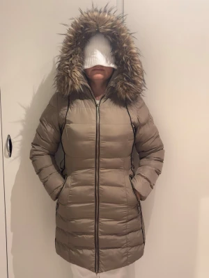 Beige lång dunjacka med fuskpäls - Snygg beige dunjacka med lång passform och huva med fuskpäls. Jackan har dragkedja framtill, två sidofickor och quiltad design. Perfekt för kalla vinterdagar och ger en stilren look.