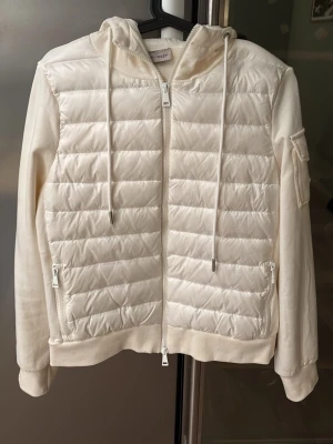 Vit cardiganjacka från Moncler - Snygg vit cardiganjacka från Moncler med quiltad front, huva med dragsko och två fickor med dragkedja. Ärmar och rygg i mjukt tyg, samt en ficka med Moncler-logga på ärmen. Perfekt för lager på lager och streetstyle.