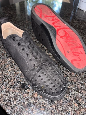 Svarta sneakers med nitar och röd sula - VILLIG ATT DISKUTERA PRIS!Säljer ett par svarta sneakers i mocka med coola svarta nitar på tån och klassisk röd sula. Skorna har svarta skosnören och en låg profil. Perfekta för dig som vill sticka ut med en edgy stil.