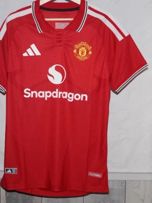 Manchester United matchtröja röd - Snygg röd Manchester United matchtröja från Adidas med klubbmärke på bröstet och Snapdragon som sponsortryck. Tröjan har vita detaljer på axlar och krage samt svartvita ränder på kragen och ärmsluten. Tillverkad i lätt och ventilerande funktionsmaterial. Är du efter en annan storlek är det bara att skriva!