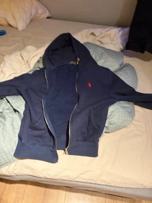 Mörkblå hoodie med dragkedja - Snygg mörkblå hoodie från Polo Ralph Lauren med dragkedja framtill och klassisk röd logga broderad på bröstet. Hoodien har huva, fickor på sidorna och ribbade muddar vid ärmslut och nederkant. Perfekt för en avslappnad och stilren look.