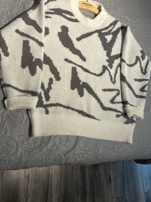 Vit stickad tröja med grått mönster - Säljer en snygg vit stickad tröja med abstrakt grått mönster. Tröjan har rund halsringning, ribbade muddar och avslut. Perfekt för dig som gillar en unik och trendig stil.