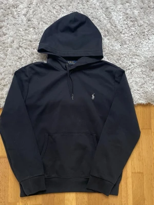 Svart hoodie från Polo Ralph Lauren - Klassisk svart hoodie från Polo Ralph Lauren med broderad logga på bröstet. Hoodien har huva med dragsko, magficka och ribbade muddar vid ärmar och nederkant. Tillverkad i mjukt bomullsmaterial som är skönt att bära.