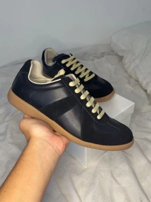 Maison margiela - Snygga svarta sneakers med beige sula och kontrasterande beige snörning. Ovandelen är i en mix av mocka och slätt läder, med en klassisk låg siluett och diskreta detaljer. Insidan är ljus och ger en fräsch look till skon.
