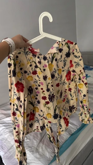 Blommig blus med knytband - Säljer en beige blus med färgglatt blommönster i rött, gult, lila och blått. Blusen har långa ärmar och knytband i midjan för en snygg siluett. Materialet känns lätt och mjukt, perfekt för vår och sommar. Rund halsringning och avslappnad passform.