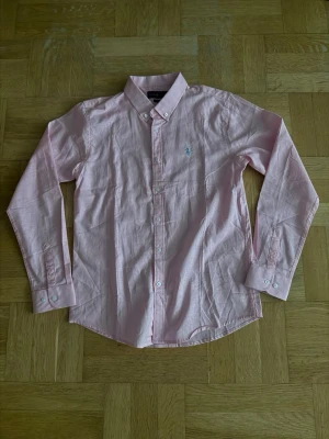 Ljusrosa skjorta från Ralph Lauren - Snygg ljusrosa skjorta från Ralph Lauren med klassisk krage och knappar framtill. Skjortan har lång ärm och den ikoniska broderade loggan på bröstet. Perfekt för en stilren look.