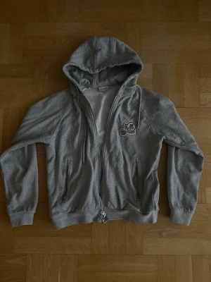 Grå hoodie från Moncler med dragkedja - Snygg grå hoodie från Moncler med dragkedja framtill och två fickor med dragkedja. Hoodien har broderade Moncler-loggor på bröstet och en mysig insida. Perfekt för en chill och stilren look.