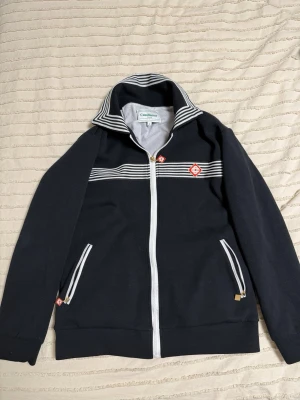 Casablanca kofta - Snygg svart hoodie från Casablanca med vita ränder över bröstet och vita dragkedjor. Har två fickor framtill och ett rött broderat logomärke på bröstet. Ståkrage och sportig design, perfekt för en trendig streetwear-look.