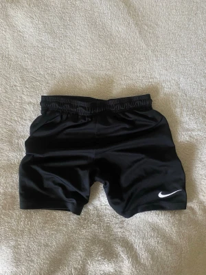 Svarta träningsshorts från Nike - Svarta sportshorts från Nike med elastisk midja och vit logga på ena benet. Tillverkade i ett glansigt, lätt material som passar perfekt för träning eller sportiga aktiviteter.