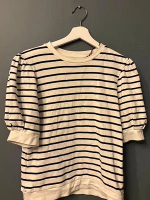 Randig topp från Holly & Whyte - Säljer en vit topp med svarta ränder från Holly & Whyte by Lindex. Toppen har puffiga trekvartsärmar och rund halsringning. Tillverkad i mjuk bomull.