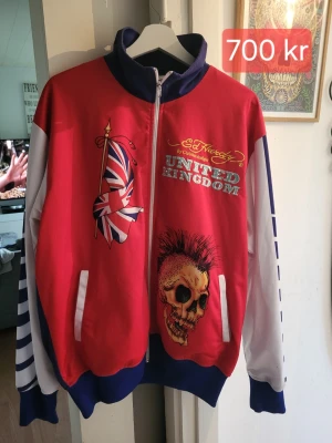 Röd Ed Hardy hoodie med tryck - Säljer en färgstark hoodie från Ed Hardy med dragkedja, stora tryck av Union Jack-flaggan och dödskalle på både fram- och baksida. Texten 'United Kingdom' och detaljer i blått, vitt och gult. Långa ärmar med blå ränder och ribbade muddar. Perfekt för dig som gillar statement-plagg.