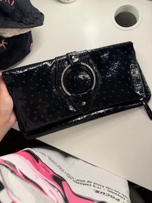 Hand väska  - Snygg svart kuvertväska i glansigt material med präglat mönster och ett stort dekorativt spänne framtill. Väskan har flera fack inuti och stängs med tryckknapp. Perfekt för att lyfta din outfit med en cool detalj.