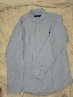 Ralph lauren skjorta - Klassisk ljusblå skjorta från Ralph Lauren i bomull. Skjortan har button-down krage, långa ärmar och knappar framtill. Ikoniska loggan broderad på bröstet. Perfekt för en stilren och avslappnad look.
