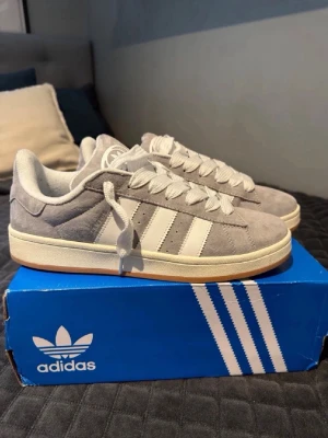 Grå Adidas sneakers i mocka - Snygga grå sneakers från Adidas med klassiska vita ränder på sidan och vit sula. Skorna är låga och har en ovandel i mjuk mocka samt vita skosnören. Perfekta för en clean och stilren look.