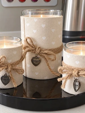Set med tre dekorerade ljuslyktor - Säljer ett set med tre glasljuslyktor klädda i beige tyg med vita hjärtan. Varje lykta är dekorerad med ett grovt snöre och ett hjärtformat metallhänge med texten 'Ich liebe Dich'. Perfekt som mysig inredningsdetalj.