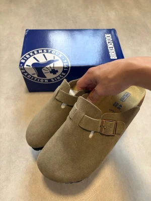 Birkenstock Boston Beige Mocka Strl 40 - Säljer ett par snygga beige Birkenstock Boston i storlek 40. Endast provade en gång inomhus och är i mycket fint skick utan några brister.  Klassisk och bekväm modell i mjuk mocka med justerbart metallspänne och ergonomisk fotbädd i kork. Perfekta både hemma och ute!  Vid bud eller funderingar är det bara att skriva! 😊