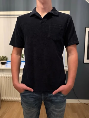 Mörkblå piké med krage och bröstficka - Snygg mörkblå t-shirt från East West med klassisk krage och bröstficka. Tillverkad i ett mjukt, frottéliknande material som ger en unik känsla. Perfekt för dig som vill ha en stilren men ändå avslappnad look.