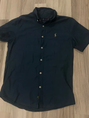 Mörkblå kortärmad skjorta Polo Ralph Lauren - Snygg mörkblå skjorta från Polo Ralph Lauren med korta ärmar och klassisk knappslå. Skjortan har den ikoniska broderade loggan på bröstet och är tillverkad i mjuk bomull. Perfekt till jeans eller chinos för en clean look.