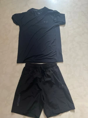 Svart träningsset från Under Armour - Säljer ett svart träningsset från Under Armour med en kortärmad t-shirt och matchande shorts. Båda plaggen är tillverkade i ett lätt och snabbtorkande material, perfekt för träning eller gym. Diskret logga på både tröja och shorts. Storlek M helt nya och går att diskutera pris