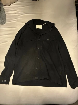 Svart overshirt från Jack & Jones - Säljer en svart overshirt från Jack & Jones i storlek M. Skjortan har klassisk krage, knäppning framtill, två bröstfickor med lock och knappar samt långa ärmar med knapp vid manschetten. Perfekt att bära som lager över t-shirt.