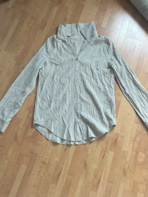 Beige randig linne skjorta med hög krage - Säljer en beige skjorta med tunna vertikala ränder och hög krage. Skjortan har lång ärm, knappar framtill och är tillverkad i ett lätt, skrynkligt tyg som ger en avslappnad känsla. Den är i väldigt bra skick och så fin att styla! Den har också lite oversized passform