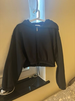 Svart cropped hoodie från FB Sister - Säljer en svart cropped hoodie från FB Sister med dragkedja framtill och huva. Tröjan har långa ärmar, resår i midjan och en avslappnad passform. Perfekt för en chill och trendig look.