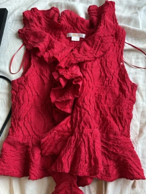 Röd volang topp från Gina Tricot - Säljer en röd ärmlös topp från Gina Tricot med volangdetaljer och struktur i tyget. Toppen har en snygg krage och omlottkänsla framtill. Perfekt för dig som vill sticka ut med en färgstark och unik look.