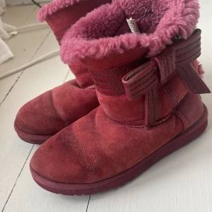 UGGs rosa storlek 37 - Rosa UGGs med rosett i sidan. Härlig varm rosa päls på insidan. Skaft i häl på ena skon är tradigt men inget som påverkar skon, en skomakare kan säkert sy fast den. 