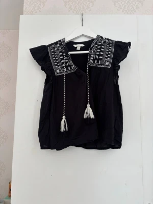 Svart blus med broderi och knytband  - Svart blus med vita broderade detaljer på axlarna och v-ringning med dekorativa knytband med tofsar. Blusen har volangärmar och en lös passform som ger en avslappnad look. Perfekt för dig som gillar boho-stil.