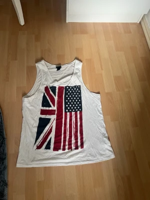 Vit linnetopp med flaggtryck - Säljer en vit linnetopp med tryck av brittiska och amerikanska flaggan på framsidan. Toppen är ärmlös och har en avslappnad passform, perfekt för varma dagar. Materialet känns mjukt och lätt mot huden.