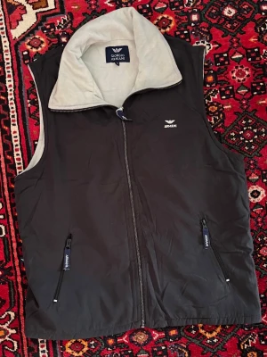 Svart dunväst från Armani - Svart dunväst från Armani med grå insida och hög krage. Västen har dragkedja framtill och två fickor med dragkedja. Diskret Armani-logga på bröstet och detaljer i blått på dragkedjorna. Perfekt för lager-på-lager och stilren look.