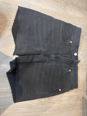 Svarta jeansshorts - Säljer ett par svarta jeansshorts med rå, avklippt kant och klassisk femficksdesign. Shortsen har hög midja och stängs med knapp och dragkedja. Perfekta för sommaren och ger en avslappnad look. Helt oanvända