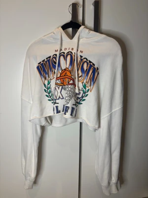 Vit cropped hoodie - Säljer en vit cropped hoodie med huva och dragsnören. Framsidan har ett stort färgglatt tryck med texten 'Wisconsin' och en basketkorg. Perfekt för en sportig och avslappnad stil. Använd fåtal gånger men är i gott skick