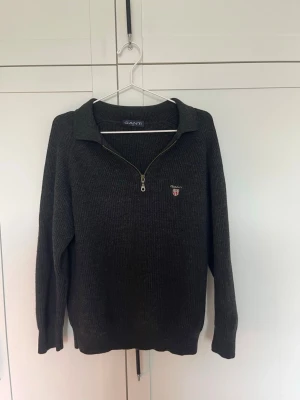 Mörkgrå stickad tröja från Gant - Mörkgrå stickad vintage tröja från Gant med halv dragkedja och krage. Tröjan har ribbad struktur och ett broderat Gant-emblem på bröstet. Perfekt för lager på lager och en stilren look.