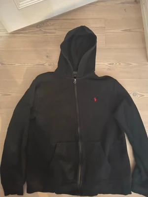 Svart hoodie med dragkedja - Säljer en svart hoodie med dragkedja och huva. Klassisk design med känguruficka framtill och en liten röd logga broderad på bröstet. Perfekt för en chill och avslappnad stil. Passar till det mesta och är riktigt bekväm.