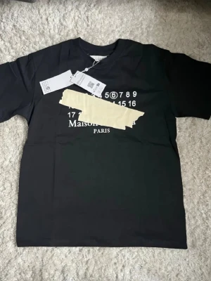 Svart t-shirt med tryck Maison Margiela - Svart t-shirt med Maison Margiela-tryck och grafisk tejpdetalj över bröstet. Klassisk passform och rund hals. T-shirten har korta ärmar och är tillverkad i mjuk bomull. Perfekt för dig som gillar streetwear och unika prints. Inkluderar dustbag från masion margiela 