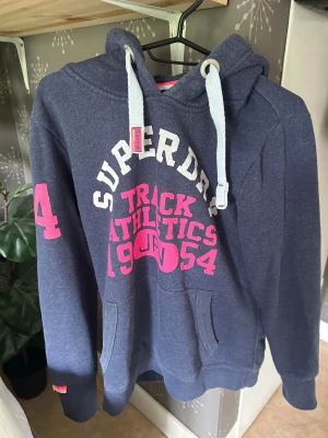 Blå Superdry hoodie med tryck - Mörkblå hoodie från Superdry, den är köpt på Sellpy men jag har aldrig använt den. Den är i bra skick men texten har spruckit lite. Den är i storlek XL men sitter som en S/M.
