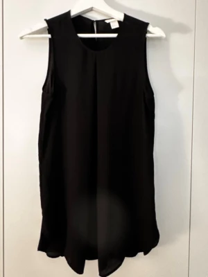 Svart ärmlös blus med rund hals - Stilren svart blus i luftig och lätt material med rund halsringning och ärmlös design. Blusen har en rak passform och något längre längd, vilket ger en snygg och avslappnad look. Perfekt att matcha med jeans eller kjol.