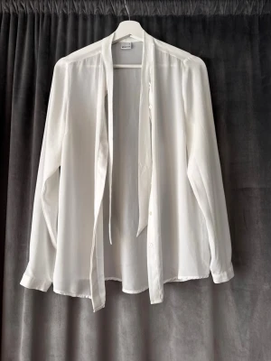 Vit blus med knytband från Gina Tricot - Säljer en stilren vit blus från Gina Tricot i storlek 34. Blusen har långa ärmar, knappar framtill och ett snyggt knytband vid kragen. Tillverkad i ett lätt och luftigt material som ger en avslappnad men chic look. Aldrig använd!