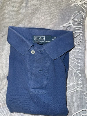 Marinblå pikétröja från Ralph Lauren - Klassisk marinblå pikétröja från Ralph Lauren med krage, knappar framtill och den ikoniska broderade loggan på bröstet. Tillverkad i mjuk bomull och har korta ärmar. Perfekt för en clean och stilren look.