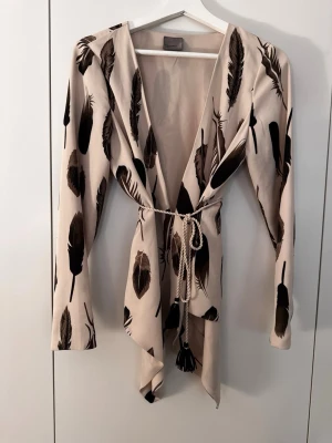 Beige tunika med fjädermönster - Beige kappa med svartbrunt fjädermönster och långa ärmar. Kappan har ett knytband i midjan med dekorativa tofsar och en öppen, avslappnad passform. Materialet ser ut att vara mjukt och följsamt, perfekt för dig som gillar unika detaljer.