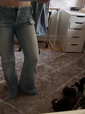 Ljusblå bootcut jeans med fickdetaljer - Säljer ett par ljusblå bootcut jeans med hög midja och unika fickdetaljer framtill. Jeansen har en klassisk dragkedja och knappstängning samt utsvängda ben. Perfekta för dig som gillar retrovibbar och en avslappnad stil.
