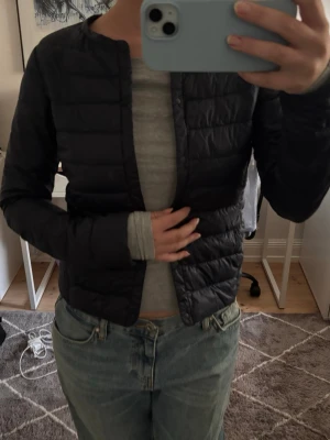Svart tunn pufferjacka - Säljer en svart, tunn pufferjacka med rund halsringning och diskreta horisontella sömmar. Jackan har en minimalistisk look utan synliga loggor eller detaljer och stängs med tryckknappar framtill. Perfekt för lager-på-lager under kalla dagar.