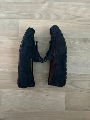 Mörkblå mocka loafers med snörning - Snygga mörkblå loafers i mocka med dekorativ snörning framtill och robust gummisula med mönster för extra grepp. Skorna har en klassisk, stilren form och passar perfekt till jeans eller chinos för en avslappnad look. De är i stort sätt oanvända.