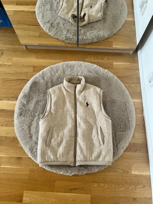 Polo Ralph lauren Beige stickad väst med dragkedja - Polo Ralph lauren Snygg beige väst med stickat kabelmönster och hög krage. Västen har två fickor framtill och stängs med dragkedja. Perfekt för lager-på-lager och ger en chill men stilren look.
